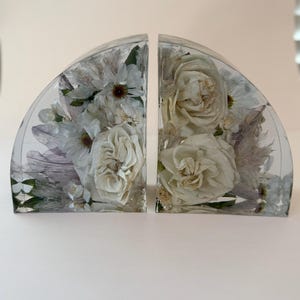 May include: Two clear, semi-circular bookends with white flowers encased in resin. The bookends feature roses and other delicate blooms, creating a decorative and artistic display. The resin is transparent, allowing the flowers to be fully visible.
