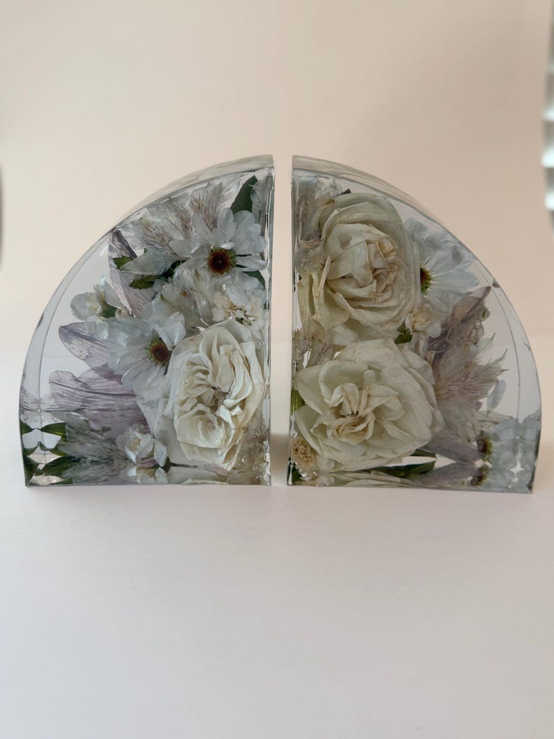 May include: Two clear, semi-circular bookends with white flowers encased in resin. The bookends feature roses and other delicate blooms, creating a decorative and artistic display. The resin is transparent, allowing the flowers to be fully visible.