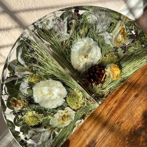 May include: A half-circle resin piece with embedded dried flowers, pine needles, and a small pine cone. The resin is clear, showcasing the natural elements. The other half is a wooden surface.