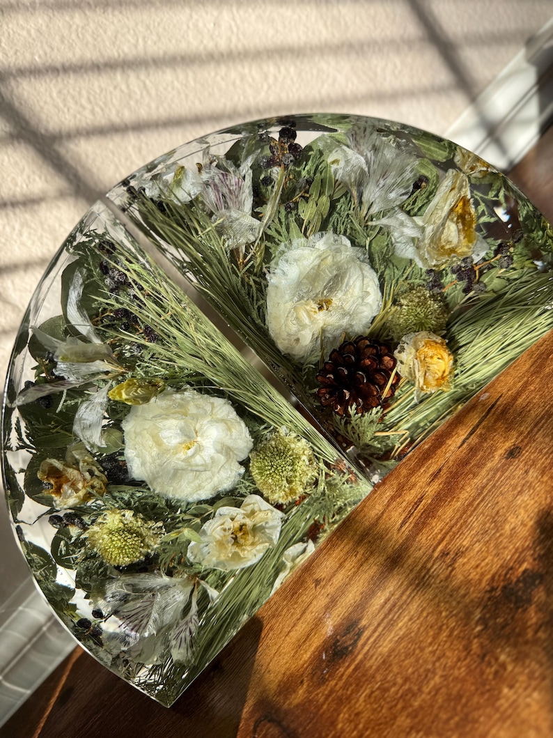 May include: A half-circle resin piece with embedded dried flowers, pine needles, and a small pine cone. The resin is clear, showcasing the natural elements. The other half is a wooden surface.