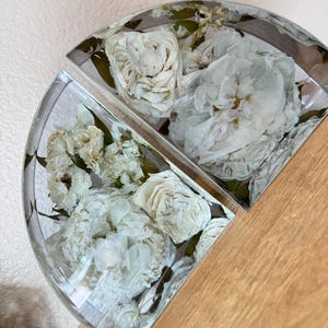 May include: Two half-circle resin pieces, each encasing white roses and green leaves. The clear resin allows a full view of the preserved flowers. The pieces are displayed on a wooden surface.