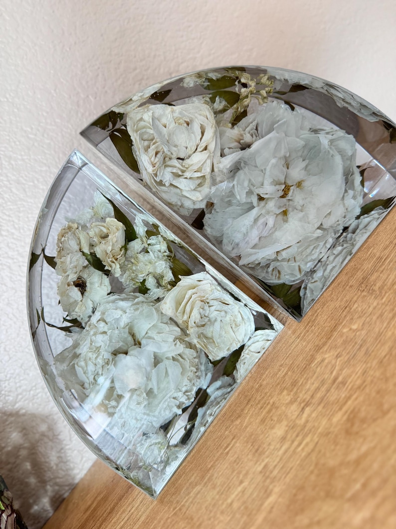 May include: Two half-circle resin pieces, each encasing white roses and green leaves. The clear resin allows a full view of the preserved flowers. The pieces are displayed on a wooden surface.