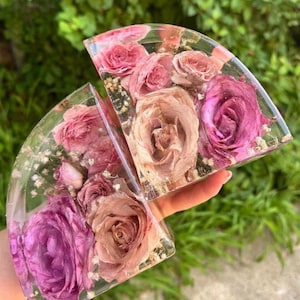 May include: Two semi-circular resin pieces, each embedded with dried roses in shades of pink and mauve. The clear resin allows the flowers to be fully visible, creating a unique decorative item. The pieces are held up against a green background.