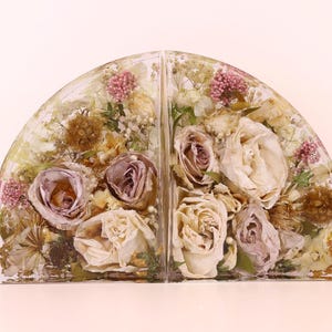 May include: Two resin bookends, each in a semi-circular shape, encasing dried flowers. The flowers include roses in shades of cream and mauve, with accents of pink and brown. The bookends are a decorative item.