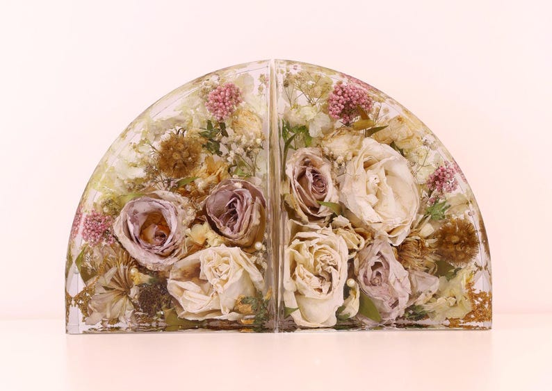 May include: Two resin bookends, each in a semi-circular shape, encasing dried flowers. The flowers include roses in shades of cream and mauve, with accents of pink and brown. The bookends are a decorative item.