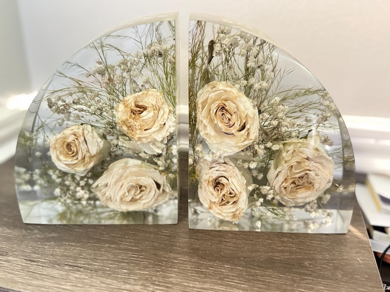 May include: Two clear, semi-circular bookends with dried cream-colored roses and white filler flowers encased in resin. The bookends are displayed on a wooden surface, showcasing a decorative floral arrangement.