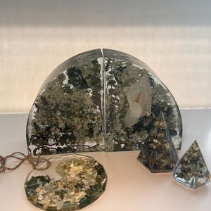 May include: A collection of clear resin objects with dried flowers and foliage. Includes a set of bookends, a round ornament, and two pyramid-shaped paperweights. The bookends are arched and filled with white flowers and greenery.