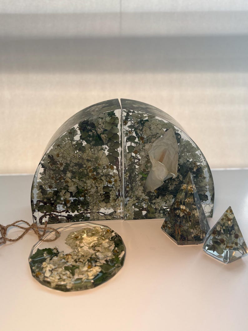 May include: A collection of clear resin objects with dried flowers and foliage. Includes a set of bookends, a round ornament, and two pyramid-shaped paperweights. The bookends are arched and filled with white flowers and greenery.