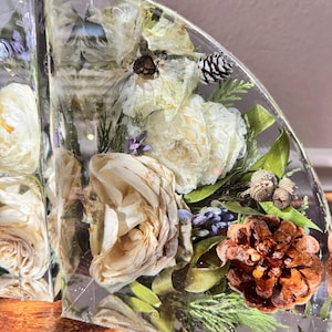 May include: Clear resin bookends with dried flowers, pinecones, and greenery encased within. The bookends feature white and cream-colored flowers, green leaves, and small pinecones, creating a natural, decorative accent for any bookshelf.