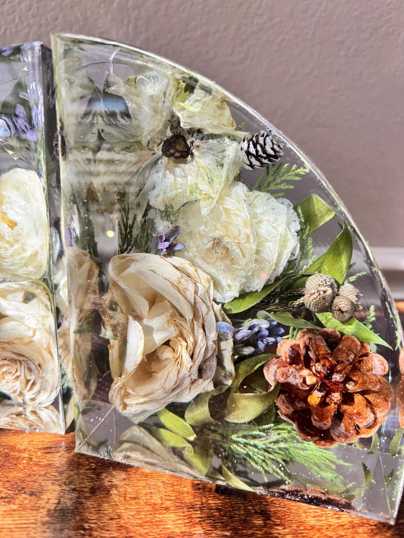 May include: Clear resin bookends with dried flowers, pinecones, and greenery encased within. The bookends feature white and cream-colored flowers, green leaves, and small pinecones, creating a natural, decorative accent for any bookshelf.
