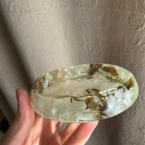 May include: A clear, oval-shaped resin dish with embedded white and brown dried flowers. The dish has a shallow, rounded design, showcasing the natural elements encased within the transparent material. The background is a light-colored fabric.