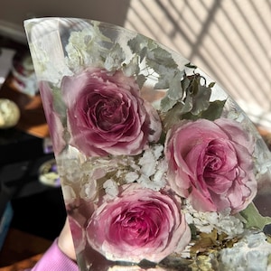 May include: A clear resin art piece featuring several pink roses and white filler flowers. The flowers are encased in a clear, curved shape, creating a unique decorative item. The roses have a deep pink hue, and the piece is well-lit.