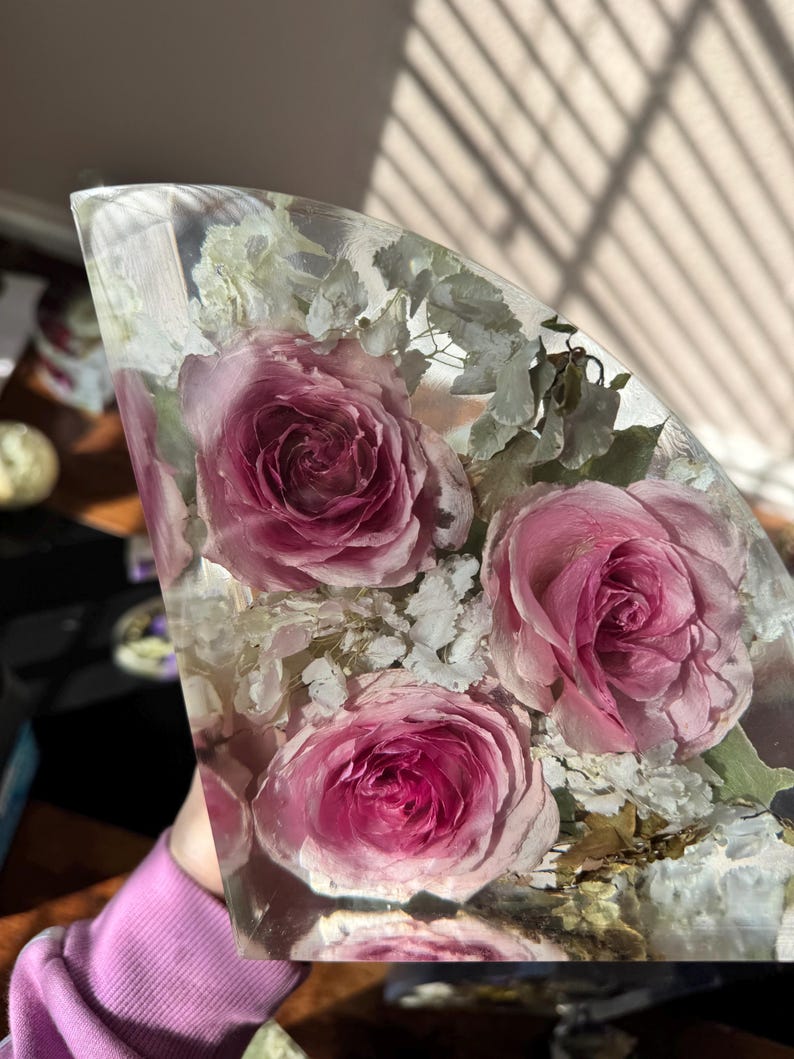 May include: A clear resin art piece featuring several pink roses and white filler flowers. The flowers are encased in a clear, curved shape, creating a unique decorative item. The roses have a deep pink hue, and the piece is well-lit.