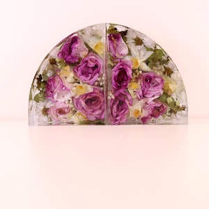 May include: Two clear, semi-circular bookends with dried flowers encased within. The flowers are primarily purple roses, with white and yellow accents. The bookends are displayed on a light pink surface.