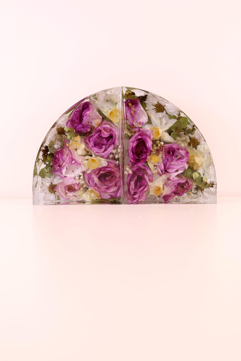 May include: Two clear, semi-circular bookends with dried flowers encased within. The flowers are primarily purple roses, with white and yellow accents. The bookends are displayed on a light pink surface.