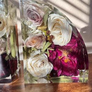 May include: Clear resin bookends with preserved flowers. One bookend features white, pink, and burgundy roses and other dried flowers. The other bookend has a similar design. The bookends are on a wooden surface.