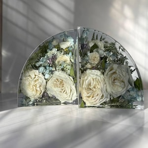 May include: Two clear, semi-circular bookends filled with preserved white roses, blue hydrangeas, and greenery. The bookends are displayed on a white surface, with shadows cast across the scene. The floral arrangement is encased in clear resin.