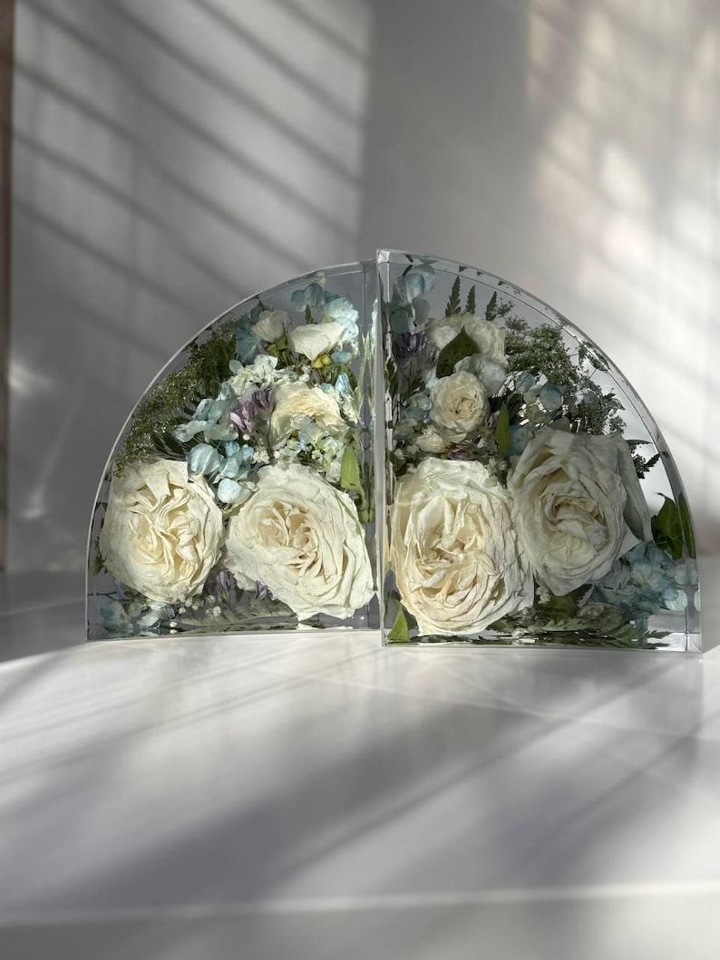 May include: Two clear, semi-circular bookends filled with preserved white roses, blue hydrangeas, and greenery. The bookends are displayed on a white surface, with shadows cast across the scene. The floral arrangement is encased in clear resin.