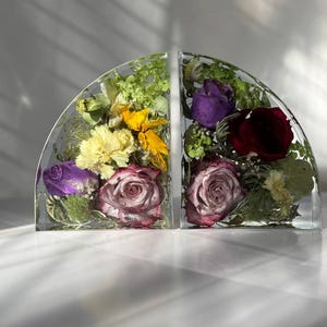 May include: Two clear, semi-circular bookends, each encasing a vibrant arrangement of dried flowers. One features yellow, purple, and pink blooms, while the other showcases deep red and purple roses. The bookends sit on a white surface.