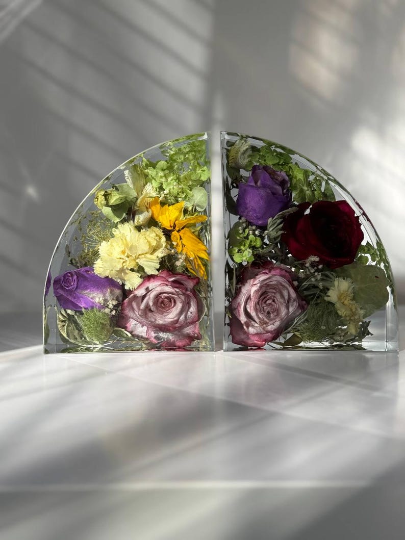 May include: Two clear, semi-circular bookends, each encasing a vibrant arrangement of dried flowers. One features yellow, purple, and pink blooms, while the other showcases deep red and purple roses. The bookends sit on a white surface.