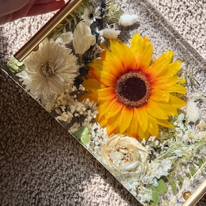 Könnte beinhalten: Ein rechteckiges Tablett aus Kunstharz mit goldenen Griffen, eingebettet mit einer leuchtend gelben Sonnenblume, weißen und cremefarbenen Blumen und Grün. Das Tablett ist ein dekorativer Artikel, perfekt zum Servieren oder Präsentieren von Gegenständen.