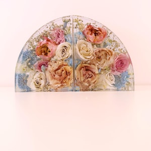 May include: A clear, arched resin piece with dried flowers. The arrangement features roses in shades of pink, peach, and cream, with blue accents. The piece is divided into two sections, creating a symmetrical display.