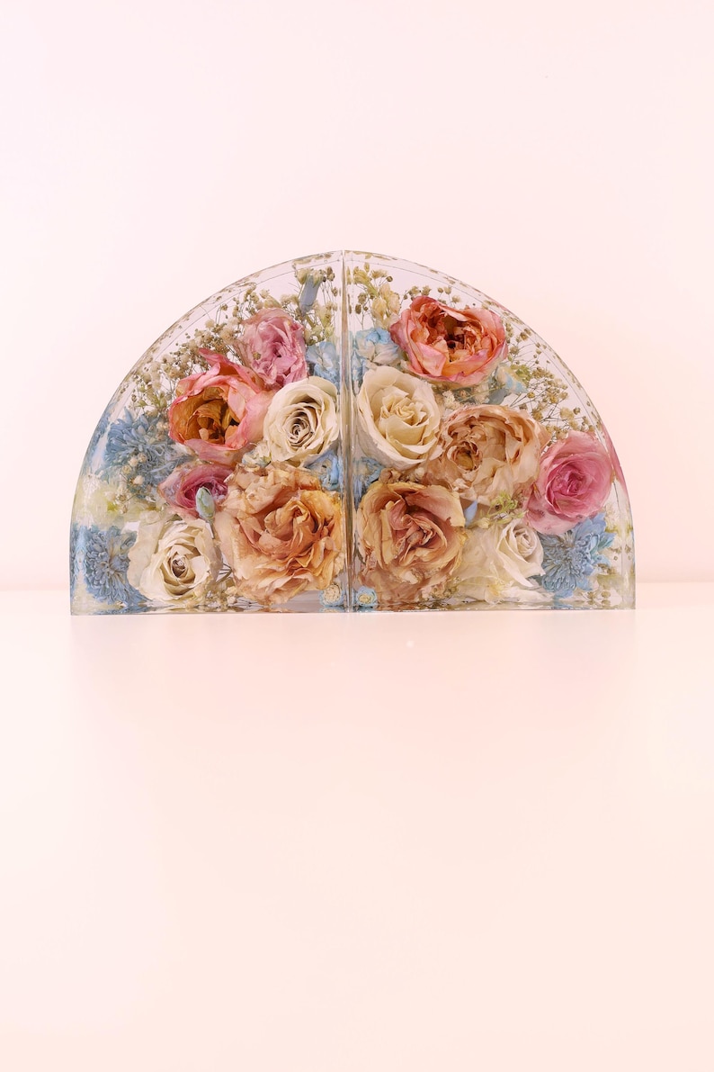 May include: A clear, arched resin piece with dried flowers. The arrangement features roses in shades of pink, peach, and cream, with blue accents. The piece is divided into two sections, creating a symmetrical display.