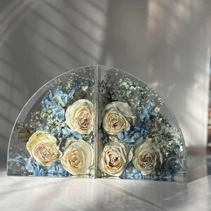 May include: Two clear, semi-circular bookends with preserved cream-colored roses and blue and white dried flowers. The bookends are made of clear resin and are displayed on a white surface.