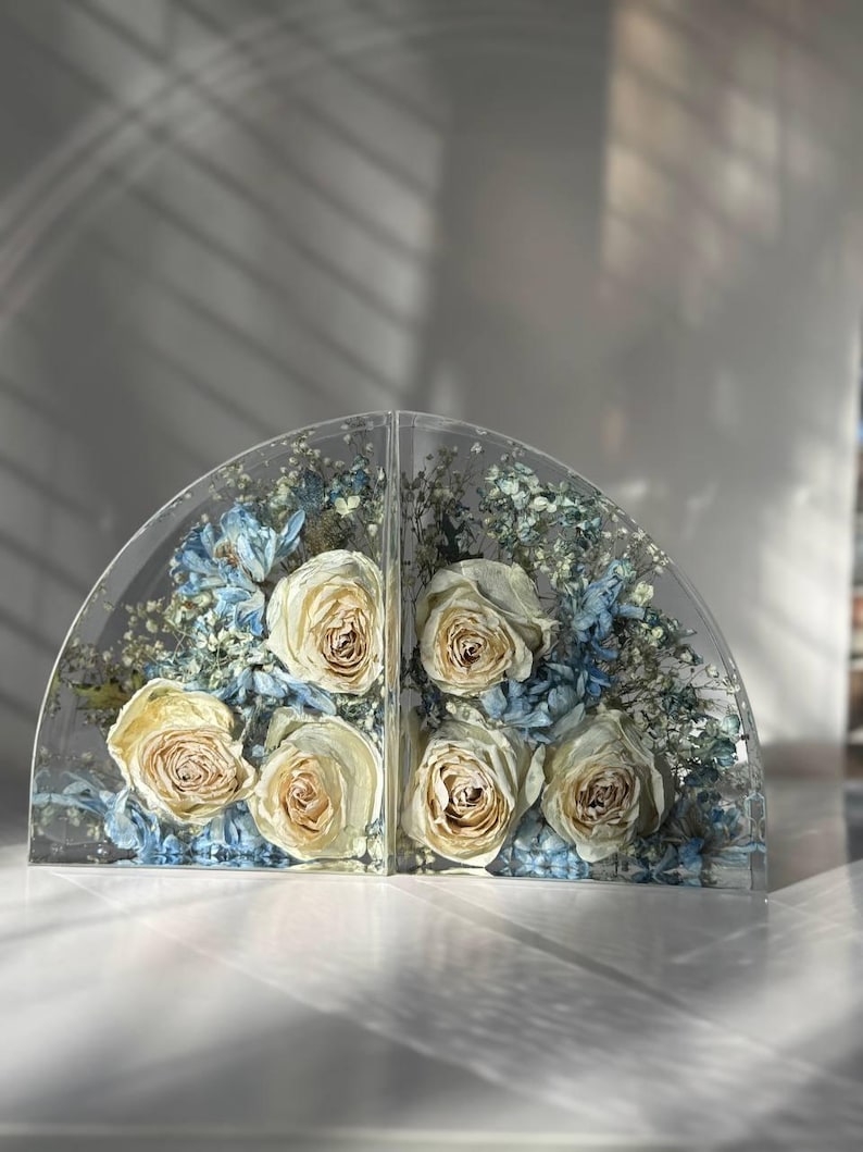 May include: Two clear, semi-circular bookends with preserved cream-colored roses and blue and white dried flowers. The bookends are made of clear resin and are displayed on a white surface.