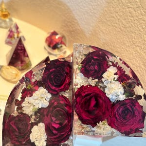 May include: Two clear, semi-circular bookends with embedded burgundy roses and white flowers. The flowers are encased in clear resin, creating a decorative and functional item. The bookends are designed to hold books upright.