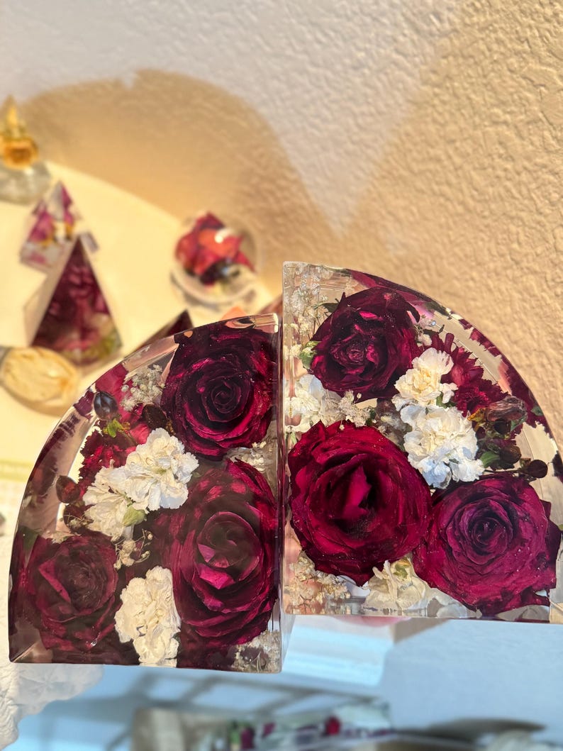 May include: Two clear, semi-circular bookends with embedded burgundy roses and white flowers. The flowers are encased in clear resin, creating a decorative and functional item. The bookends are designed to hold books upright.