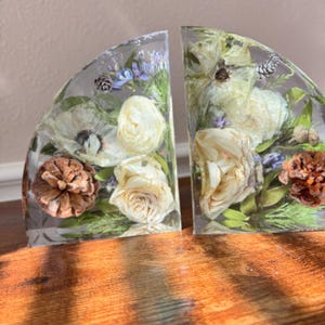 May include: Two clear resin bookends, each in a semi-circular shape, encasing dried white roses, pinecones, and greenery. The bookends are set on a wooden surface, with sunlight casting shadows across the wood.