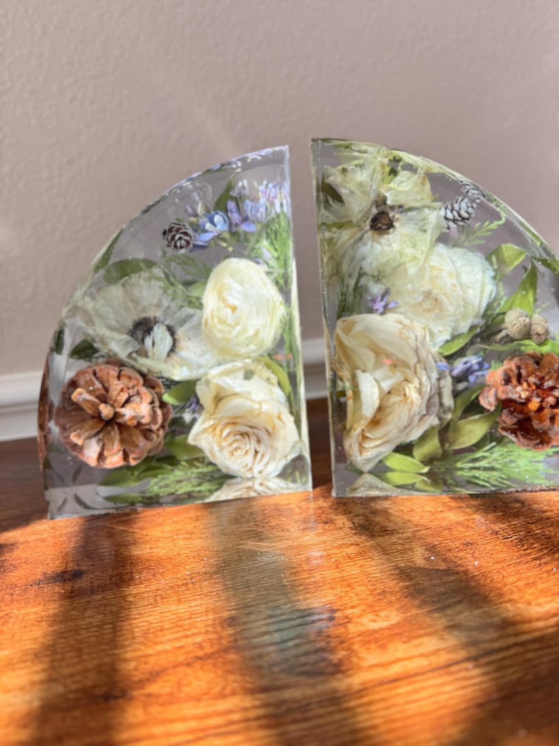 May include: Two clear resin bookends, each in a semi-circular shape, encasing dried white roses, pinecones, and greenery. The bookends are set on a wooden surface, with sunlight casting shadows across the wood.