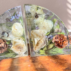 May include: Two clear, semi-circular bookends with dried flowers and pinecones encased in resin. The bookends feature cream-colored roses, lavender, and green foliage. The resin is clear, allowing the natural elements to be visible.
