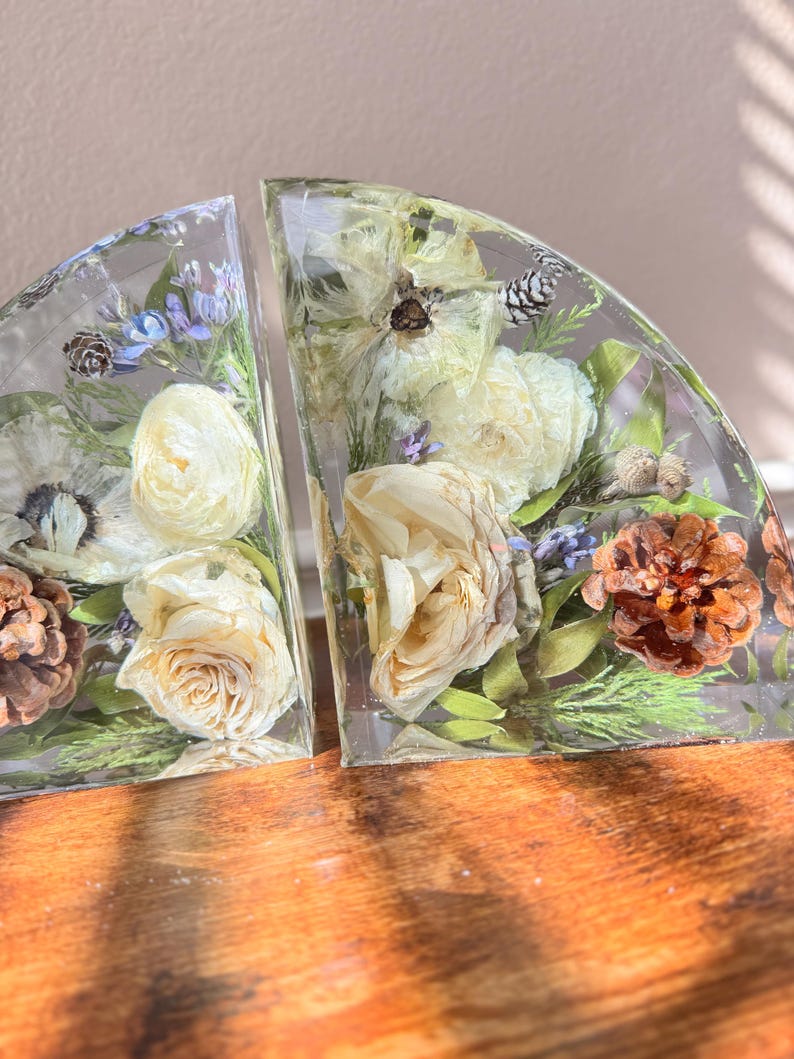 May include: Two clear, semi-circular bookends with dried flowers and pinecones encased in resin. The bookends feature cream-colored roses, lavender, and green foliage. The resin is clear, allowing the natural elements to be visible.