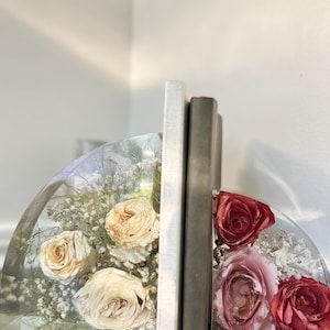 May include: Clear resin bookends with dried roses and baby's breath. One bookend features cream-colored roses, the other has pink and red roses. Two books stand between the bookends on a wooden surface.
