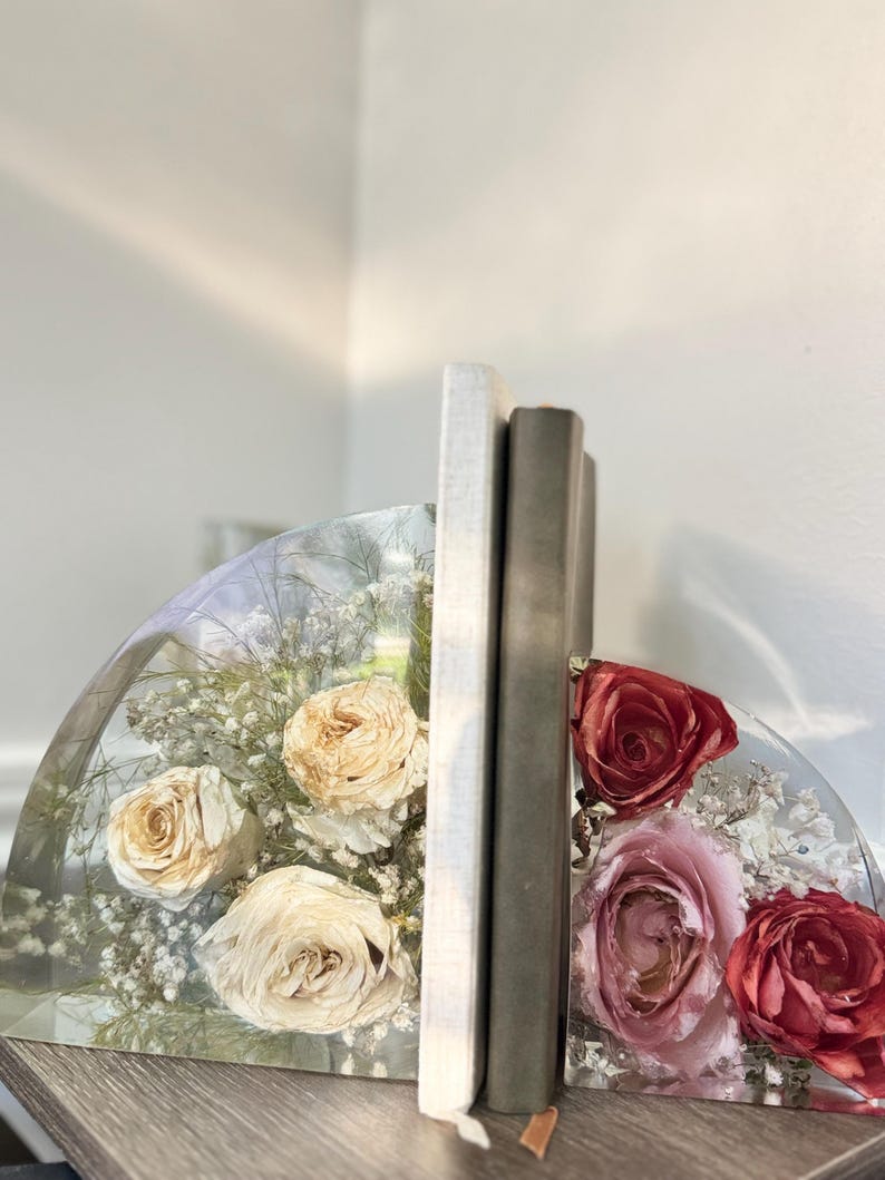 May include: Clear resin bookends with dried roses and baby's breath. One bookend features cream-colored roses, the other has pink and red roses. Two books stand between the bookends on a wooden surface.