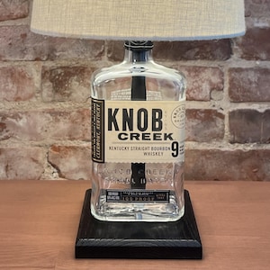 May include: A lamp made from a Knob Creek Kentucky Straight Bourbon Whiskey bottle with a beige fabric lampshade. The bottle is clear glass with a black label that reads "Knob Creek Kentucky Straight Bourbon Whiskey 9 Years Old 100 Proof". The lamp sits on a black wooden base.