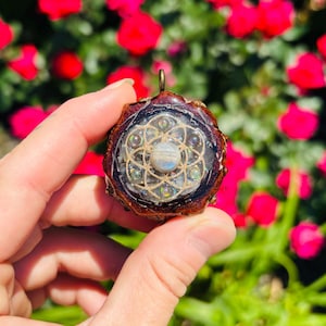 May include: A handcrafted pendant with a circular design. The pendant features a central, iridescent stone surrounded by a geometric pattern and encased in a dark brown resin. A small, gold-colored loop is at the top for hanging.