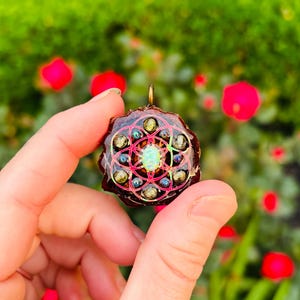 May include: A handcrafted pendant with a dark brown base, adorned with a geometric design in pink, green, and blue. The pendant features a central opal-like stone and a gold-colored bail. The background is blurred with green foliage and red flowers.