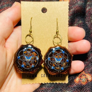 Puede incluir: Pendientes artesanales con una base marrón oscuro de forma orgánica y un diseño detallado. Cada pendiente presenta una mariposa azul y detalles azules, suspendidos de ganchos dorados. Presentados en una tarjeta beige. Aproximadamente 3 cm de diámetro.