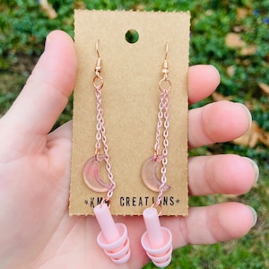 May include: A pair of pink earplugs with a crescent moon charm hanging from a gold chain. The charms are attached to the earplugs with a gold hook. The earplugs are on a brown card with the text "KM Creations" printed on it.