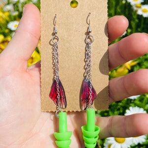 May include: A pair of earrings with a silver chain and a red butterfly charm. The earrings are attached to green silicone earplugs.