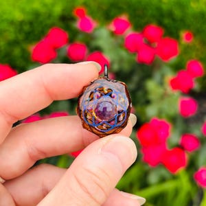 May include: A handcrafted pendant featuring a central purple gemstone, surrounded by intricate blue and gold filigree. The pendant has a unique, organic shape and is suspended from a small metal loop. The background is blurred with vibrant red flowers and green foliage.
