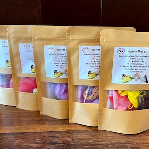 May include: Five craft kits in brown paper bags with clear windows. Each kit contains supplies for making flower dolls, including floral wire, wooden beads, artificial flowers, and embroidery floss. The kits have labels with the text "Flower Doll Kit".