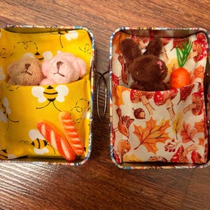 May include: Two small metal containers. One has a yellow bee-themed fabric pocket with two teddy bears and candy. The other has a fall-themed fabric pocket with a brown bunny and a carrot. Both containers have a metal handle.