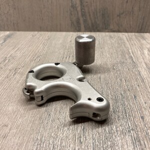 May include: A silver metal archery release aid with a black trigger and a silver knob on top. The release aid is designed to be used with a bow and arrow.