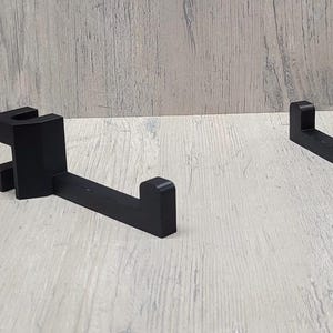 May include: Two black, right-angled brackets. Each bracket has a rectangular base with a vertical support and a horizontal arm. The brackets are made of a solid, matte black material and are designed to hold an object.