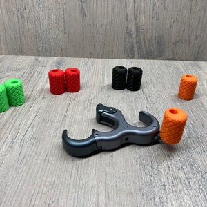 May include: A set of archery release aids with different colored rubber grips. The release aids are made of metal and have a trigger mechanism. The grips are red, green, black, and orange.