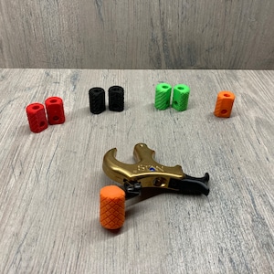 May include: A gold archery release aid with black and orange grip accessories. The release aid has the word "STAN" printed on it. There are also black, red, and green grip accessories.